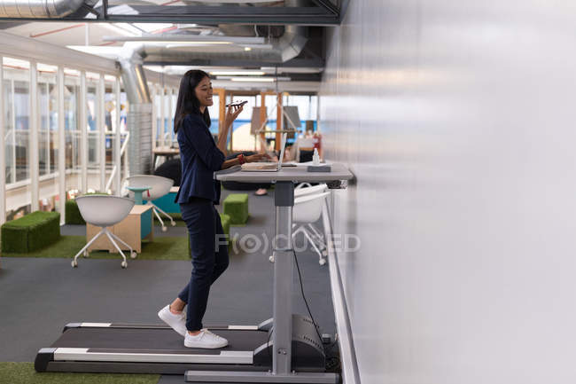 Жінка-виконавча розмова на мобільному телефоні під час тренувань на біговій доріжці в офісі — стокове фото