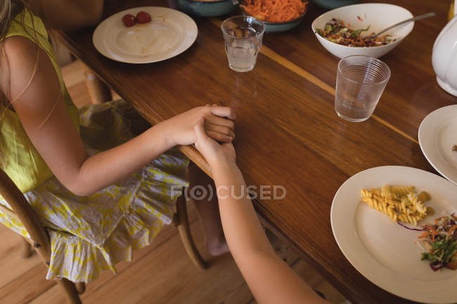 Madre cogida de la mano de su hija mientras almuerza en la cocina en casa - foto de stock
