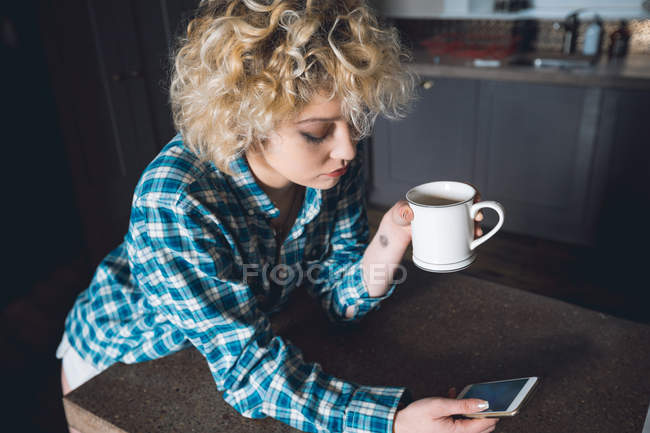 Mulher tomando café enquanto usa telefone celular na cozinha em casa — Fotografia de Stock