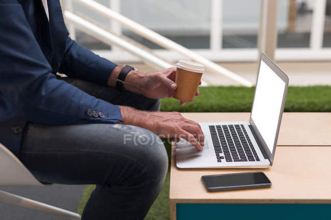 Imagen recortada de hombre de negocios tomando café mientras usa el ordenador portátil en la oficina - foto de stock