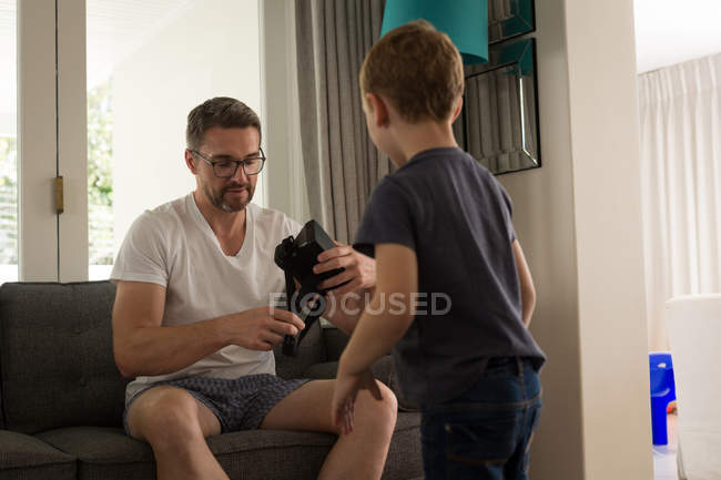 Padre con su hijo sosteniendo auriculares de realidad virtual en la sala de estar en casa - foto de stock