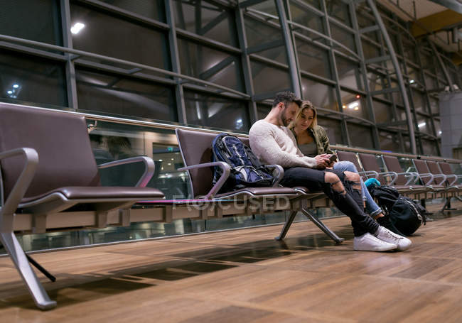 Pareja usando teléfono móvil en la sala de espera en el aeropuerto - foto de stock