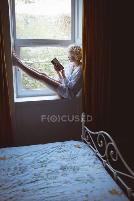 Donna che legge un libro vicino alla finestra di casa — Foto stock
