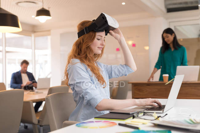 Diseñadora gráfica femenina con auriculares de realidad virtual usando portátil en la oficina - foto de stock