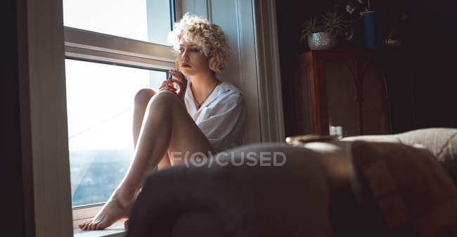 Mujer pensativa sentada cerca de la ventana en casa - foto de stock