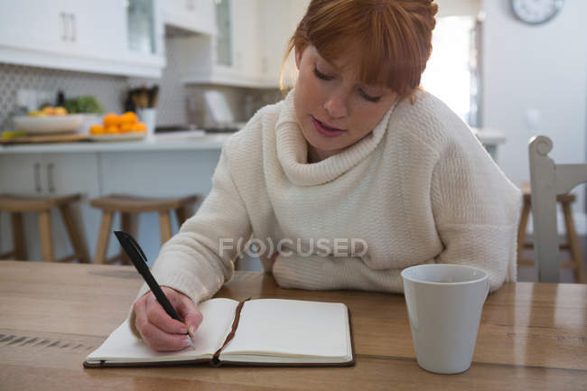 Mujer pensativa escribiendo en el diario en casa en la cocina - foto de stock