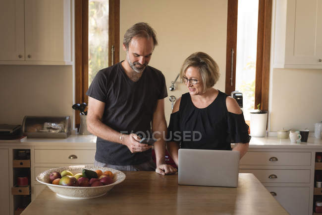 Пара с помощью мобильного телефона и ноутбука на кухне в домашних условиях — стоковое фото
