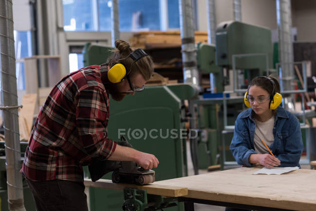 Masculino carpinteiro usando jack plano enquanto fêmea olhando para ele no workshop — Fotografia de Stock
