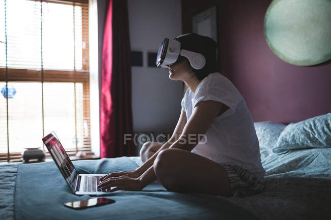 Frau benutzt Laptop mit Virtual-Reality-Headset im heimischen Schlafzimmer — Stockfoto