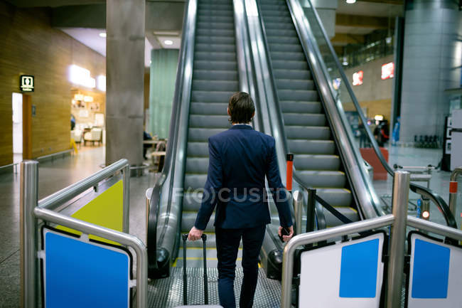 Empresario caminando con equipaje hacia escaleras mecánicas en el aeropuerto - foto de stock