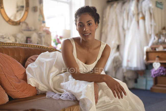 Schöne Mischlingsbraut im Brautkleid auf dem Sofa sitzend — Stockfoto