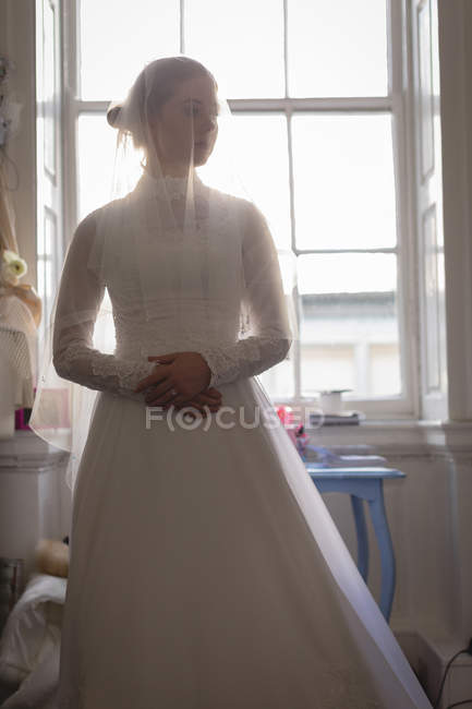 Joven novia en vestido de novia de pie cerca de la ventana en la boutique - foto de stock