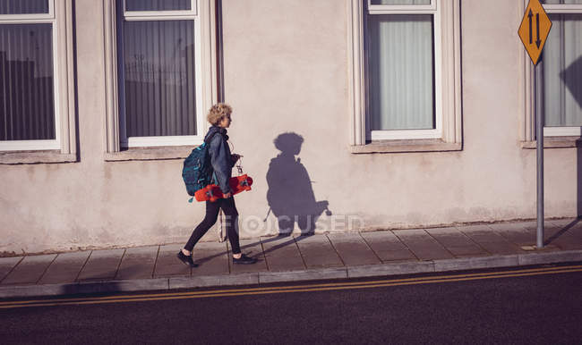 Femme marchant avec sac à dos et planche à roulettes sur un sentier dans la ville — Photo de stock