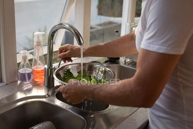 Homem sênior limpeza vegetal com água na cozinha em casa — Fotografia de Stock