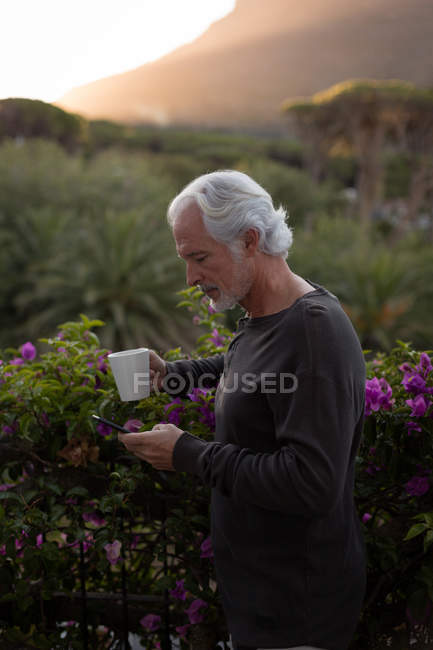 Пенсионер пьет кофе, пользуясь мобильным телефоном на балконе дома — стоковое фото