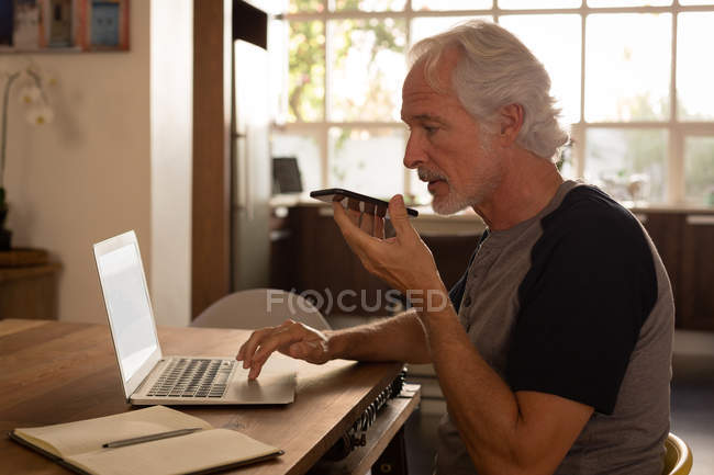 Старший чоловік розмовляє на мобільному телефоні, використовуючи ноутбук вдома — стокове фото