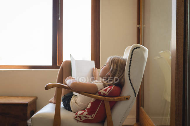 Chica estudiando en la sala de estar en casa, sentado en sillón con libro - foto de stock