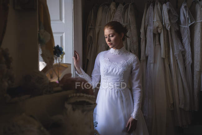 Young bride in wedding dress standing at window and thinking — Stock Photo