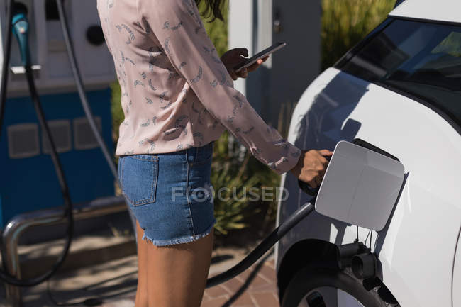Sección media de la mujer que usa el teléfono móvil mientras carga el coche eléctrico en la estación de carga - foto de stock