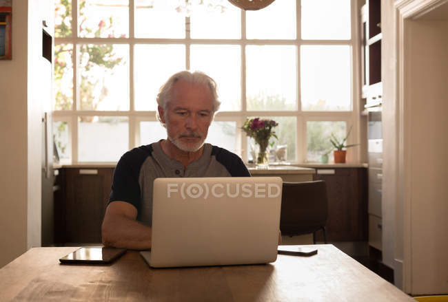 Пожилой мужчина использует ноутбук на кухне дома — стоковое фото