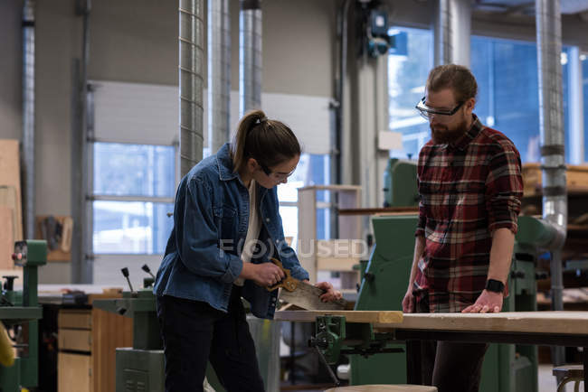 Carpinteiro feminino usando serra de mão, enquanto o macho olhando para ela na oficina — Fotografia de Stock