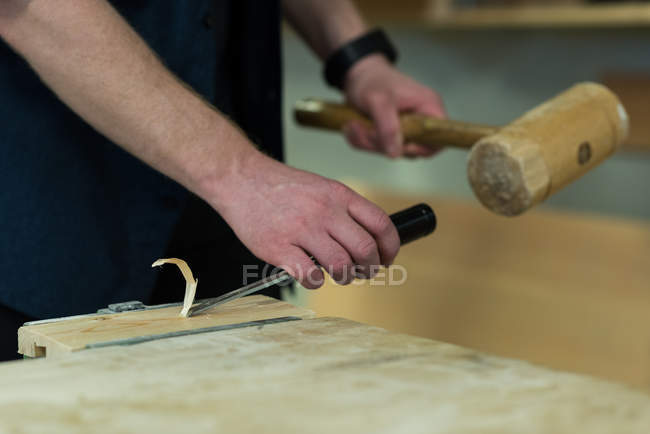Section médiane du menuisier masculin à l'aide d'un ciseau avec marteau sur un morceau de bois — Photo de stock