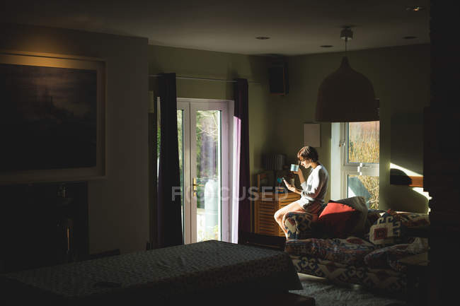 Donna che prende il caffè mentre usa il telefono cellulare in soggiorno a casa — Foto stock