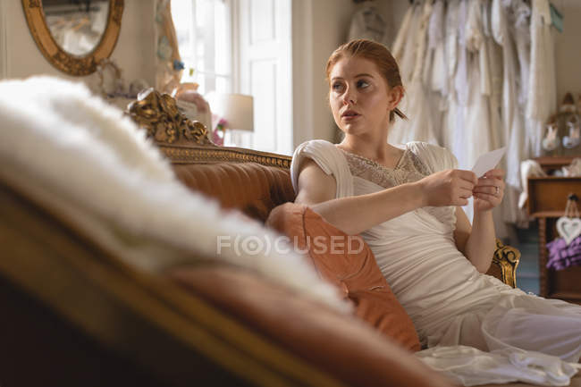 Young bride in wedding dress holding note while sitting on sofa — Stock Photo