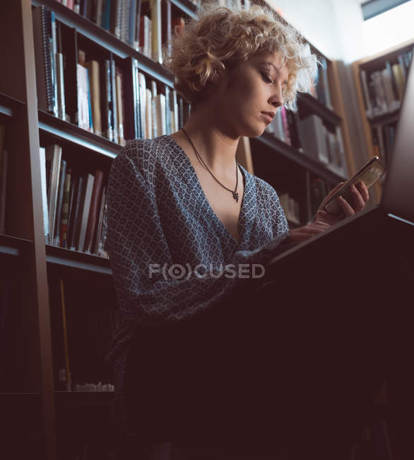 Молодая женщина с помощью мобильного телефона и ноутбука в библиотеке — стоковое фото