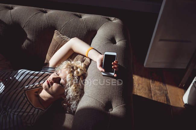 Mujer durmiendo en la sala de estar sosteniendo el teléfono móvil en casa - foto de stock