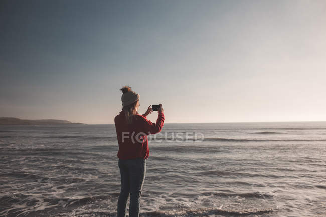 Vista trasera de la mujer tomando fotos con teléfono móvil en la playa - foto de stock