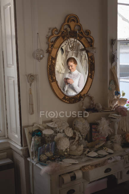 Caucasian bride in wedding dress and veil standing at mirror at vintage boutique and using mobile phone — Stock Photo