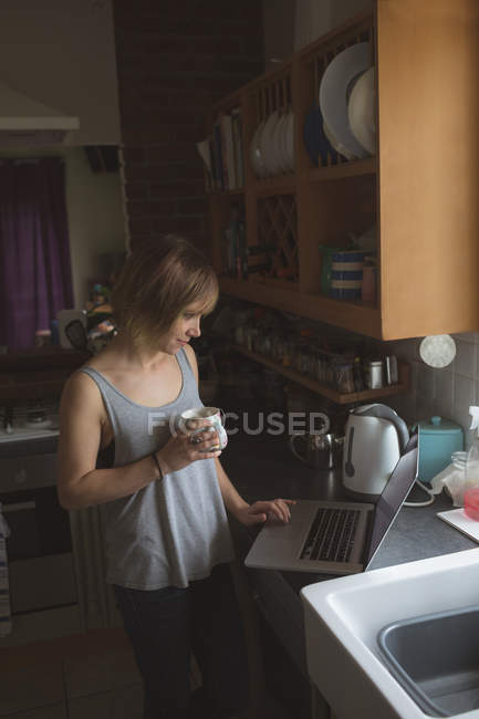 Красива жінка використовує ноутбук, маючи каву на кухні вдома — стокове фото