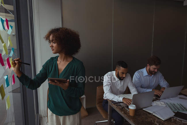 Female architect with digital tablet writing on sticky note while colleagues using laptop in the office — Stock Photo