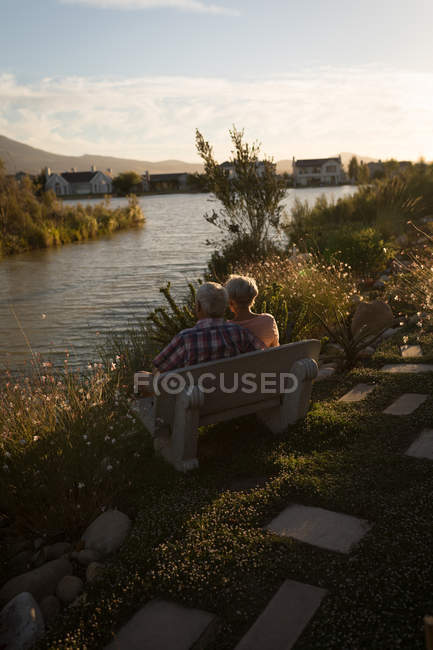 Старша пара відпочиває на лавці біля берега річки в сонячний день — стокове фото