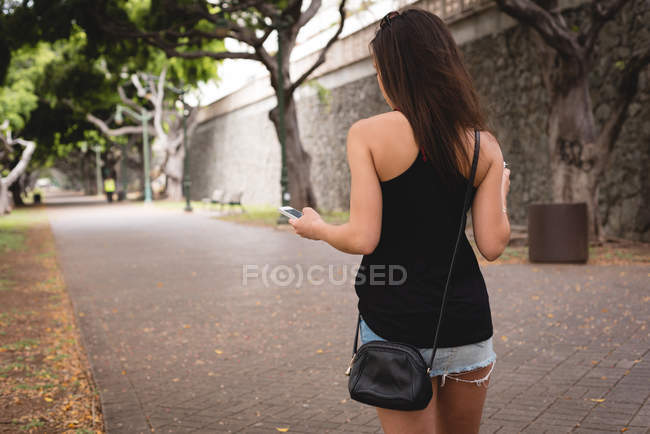 Вид ззаду жінки за допомогою мобільного телефону під час ходьби на тротуарі — стокове фото