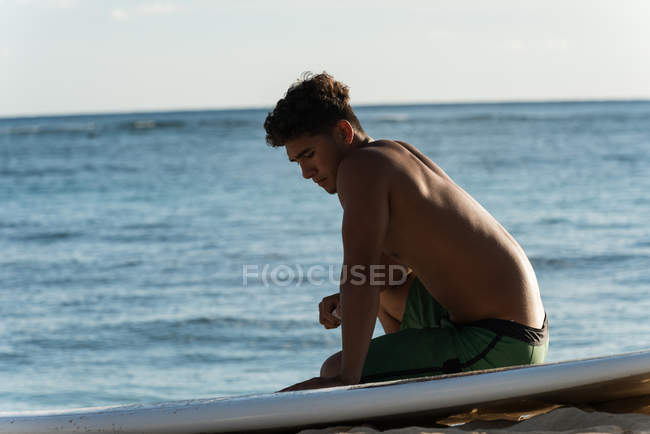 Junger männlicher Surfer sitzt mit Surfbrett am Strand — Stockfoto