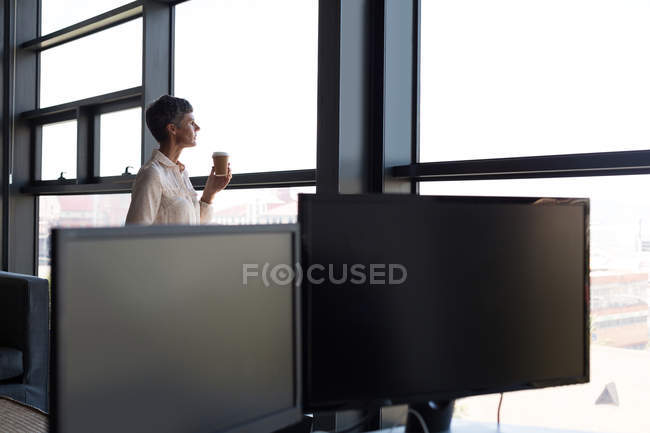 Donna d'affari matura che guarda attraverso la finestra mentre prende un caffè in ufficio — Foto stock