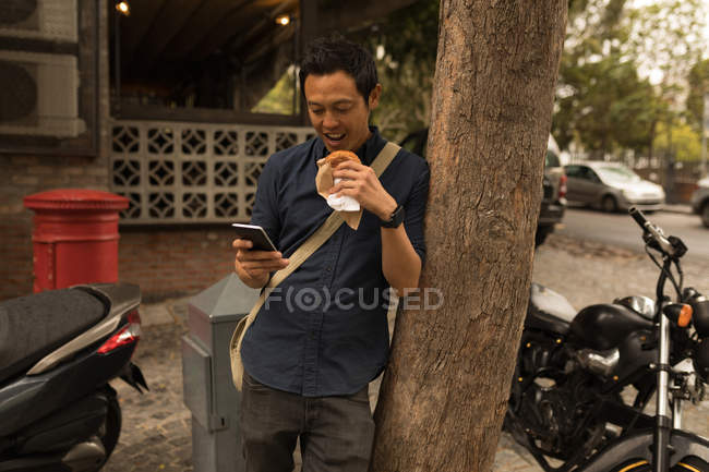 Uomo d'affari sorridente utilizzando il telefono cellulare mentre si mangia cibo — Foto stock