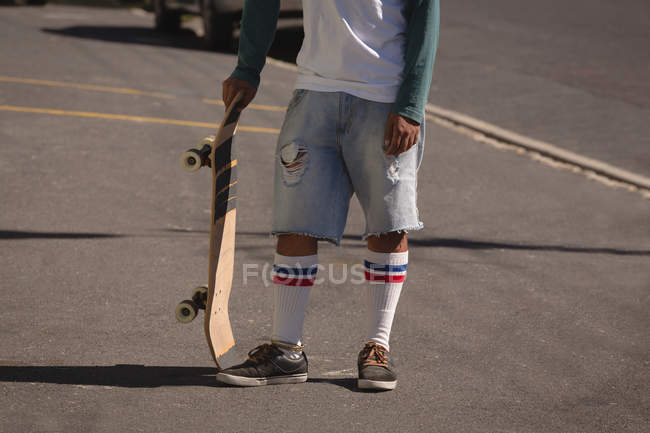 Section basse de l'homme debout avec planche à roulettes dans la rue au soleil — Photo de stock