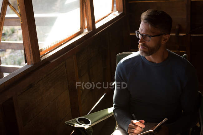 Thoughtful man sitting on chair and looking through window at log cabin — Stock Photo