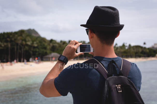 Visão traseira do homem clicando foto com telefone celular perto da praia — Fotografia de Stock