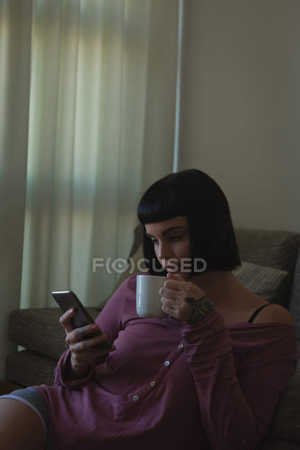 Женщина пьет кофе, используя мобильный телефон в гостиной дома — стоковое фото