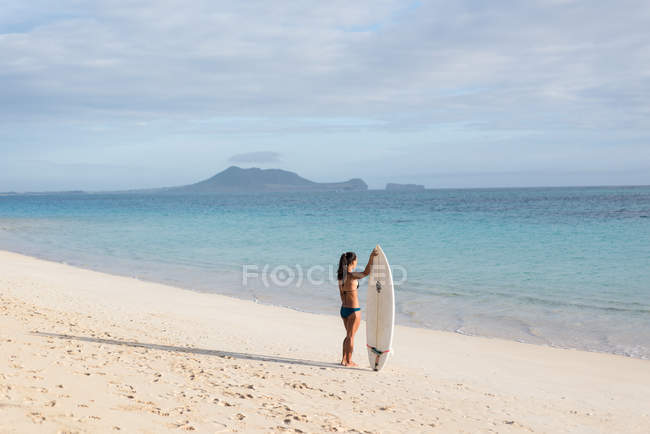 Femme debout avec planche de surf à la plage par une journée ensoleillée — Photo de stock