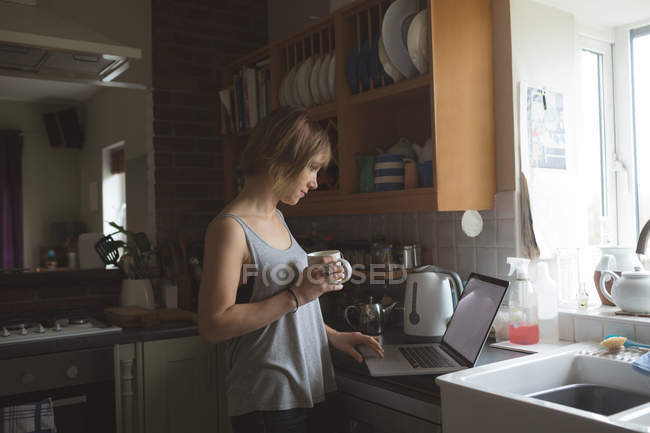 Mulher bonita usando laptop enquanto toma café na cozinha em casa — Fotografia de Stock
