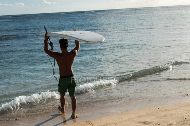 Vista traseira do surfista masculino caminhando com prancha na praia — Fotografia de Stock
