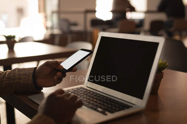Senior graphic designer using laptop and mobile phone in cafeteria at office — Stock Photo