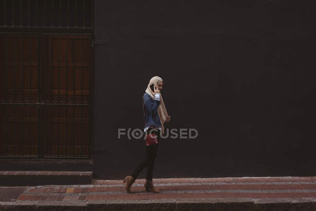Side view of hijab woman talking on mobile phone while walking on sidewalk — Stock Photo