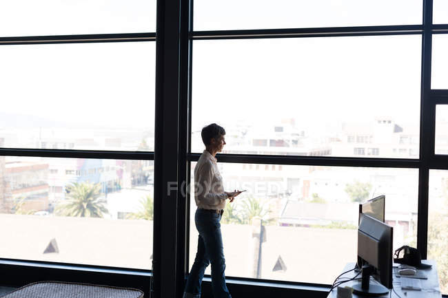 Reife Geschäftsfrau mit Handy blickt durchs Fenster ins Büro — Stockfoto