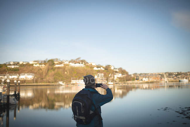 Caminhante masculino tirando foto com telefone celular perto do lago no campo — Fotografia de Stock